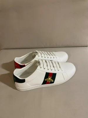 Gucci vita sneakers med bi-detalj - Snygga vita sneakers från Gucci i läder med klassiska gröna och röda ränder på sidan samt en broderad guldfärgad bi. Skorna har vita snören, svart och röd detalj på hälen och en stilren låg profil. Perfekta för dig som vill ha en ikonisk och trendig look. #gucci #gucciace #gucciacesneakers #designerskor #luxurysneakers #sneakers42 #herrsneakers #streetwearstyle #highendfashion #guccibee #vitaskor #lyxmärke #premiumskor #trendysneakers