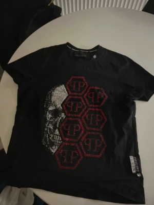 Svart Philipp Plein t-shirt med döskalle - Cool svart t-shirt från Philipp Plein med glittrande döskallemotiv och röda hexagoner med PP-logga på framsidan. Rund halsringning och korta ärmar. Märkeslapp på sidan. Perfekt för dig som gillar edgy och unik design.