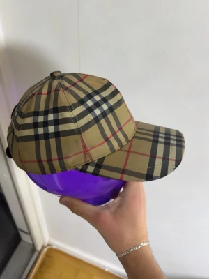 Burberry keps - Säljer en beige keps från Burberry med det ikoniska rutmönstret i svart, vitt och rött. Kepsen har böjd skärm och justerbar rem bak. Tillverkad i ett lätt och bekvämt material som passar perfekt till streetwear och trendiga outfits.