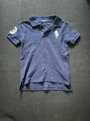 Marinblå piké från Polo Ralph Lauren - Klassisk marinblå pikétröja från Polo Ralph Lauren med vit logga på bröstet och siffran 3 på ärmen. Tröjan har korta ärmar, krage och knappar framtill. Perfekt till en avslappnad men ändå stilren look. OBS!! barn storlek