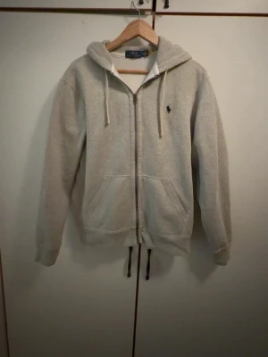 Grå hoodie från Polo Ralph Lauren - Snygg grå hoodie från Polo Ralph Lauren med dragkedja, huva och vita dragsnören. Klassisk logga broderad på bröstet. Tillverkad i mjukt bomullsmaterial och har två fickor framtill. Perfekt för en avslappnad och stilren look.