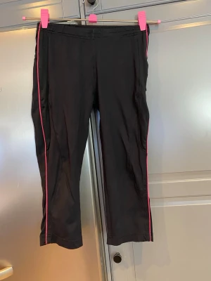 Svarta träningsbyxor med rosa detaljer strl 38 - Säljer ett par svarta träningsbyxor i caprimodell med rosa ränder längs sidorna. Byxorna har resår i midjan och är tillverkade i ett stretchigt material som passar perfekt för träning eller sportaktiviteter. Byxorna har en praktisk ficka med dragkedja på sidan.