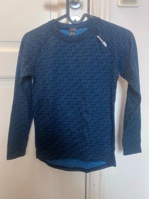 Blå underställströja från Norheim - Mönstrad blå underställströja från Norheim med långa ärmar och rund hals. Tillverkad i ett mjukt material som passar perfekt för lager-på-lager under kalla dagar. Diskret logga på bröstet och snyggt grafiskt mönster över hela tröjan. Lappen i nacken är trasig, men hänger kvar. Passar till åldern 8-10år. Säljs tillsammans med blå underställs byxor från Pierre Robert i storlek 134-140.