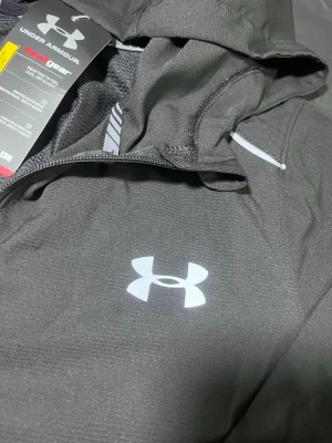 Svart vindjacka från Under Armour - Snygg svart vindjacka från Under Armour med dragkedja framtill, huva och vita detaljer. Jackan har logga på bröstet och är tillverkad i ett lätt, tekniskt material som andas. Perfekt för aktiva dagar utomhus.