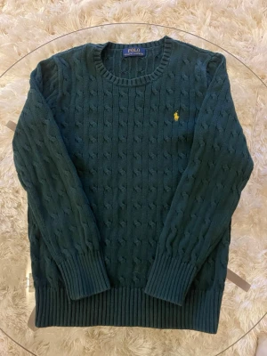 Mörkgrön kabelstickad tröja Polo Ralph Lauren - Mörkgrön kabelstickad tröja från Polo Ralph Lauren med rund halsringning och gul broderad logga på bröstet. Tröjan har långa ärmar och ribbade muddar vid ärmslut och nederkant. Perfekt för dig som gillar klassisk och stilren look.