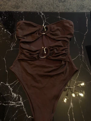 Brun baddräkt med ringdetaljer - Trendig brun baddräkt i  med två guldfärgade ringdetaljer framtill. Baddräkten har cut-out mellan byst och midja samt hög benskärning. Tillverkad i stretchigt material som passar perfekt för strand eller pool. 