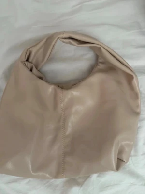 Beige slouchy handväska  - Trendig beige handväska med mjuk, slouchy form och bred axelrem. Väskan har en minimalistisk design med en synlig söm framtill. Perfekt för dig som gillar stilrena accessoarer.