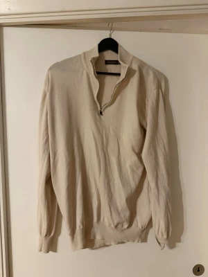 Beige halvzip tröja Oscar Jacobson - Snygg beige stickad tröja från Oscar Jacobson med halvzip och ribbade muddar. Tröjan har hög krage och är tillverkad i mjukt material som passar perfekt till lager på lager. Enkel och stilren design som funkar till många outfits. Storlek L 