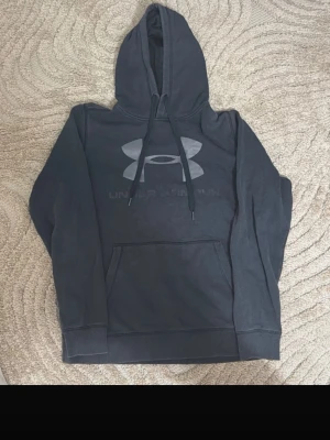 Svart hoodie från Under Armour - Svart hoodie från Under Armour med stor logga på bröstet och dragsko i huvan. Tillverkad i mjukt ColdGear-material som håller dig varm. Praktisk magficka framtill och klassisk passform, perfekt för chill dagar eller träning.