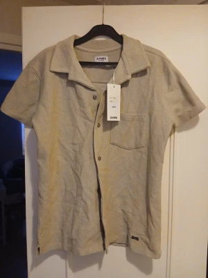 Beige skjorta från SAMS med kort ärm - Stilren beige skjorta från SAMS med korta ärmar och klassisk krage. Skjortan har knappar framtill och en bröstficka. Materialet känns mjukt och har en diskret struktur som ger en modern look. Perfekt för dig som gillar enkel och clean stil.