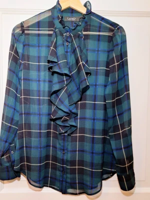 Rutig blus med volang från Lauren Ralph Lauren - Snygg rutig blus i blått, grönt och svart från Lauren Ralph Lauren. Blusen har långa ärmar, knappar framtill och en stor volang vid halsen som ger en cool statement-look. Tillverkad i ett lätt, transparent tyg som känns luftigt och trendigt. Nypris ca 2300 kr. Skick: Toppskick, finns ingenting att anmärka på utöver att tvättlappen är borttagen för att den syns annars men materialet är 100% polyester. Strl: S. Mått: • Bystvidd: 48 cm (omkrets ca 96 cm) • Axelvidd: 36 cm • Längd från axel: 66 cm