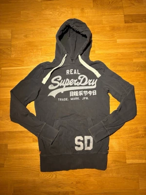 Superdry hoodie - Snygg mörkgrå hoodie från Superdry med stor vit logga och japansk text på bröstet. Storlek S och har inga defekter. Hör av dig vid frågor eller prisförslag.