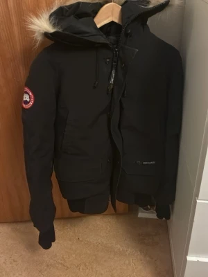 Svart parkas från Canada Goose - Säljer en svart parkasjacka från Canada Goose med avtagbar huva och äkta pälsdetalj. Jackan har ribbade muddar, två stora fickor framtill och klassisk logga på ärmen. Perfekt för kalla dagar och riktigt snygg streetstil.