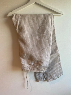 Beige randiga linnebyxor H&M 38 - Snygga beige linnebyxor med vita tunna ränder och avslappnad passform. Byxorna har resår och dragsko i midjan för extra komfort. Perfekta för en stilren look. Tillverkade i svalt och luftigt linne.