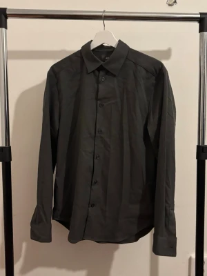 Grå skjorta från H&M - Mörkgrå skjorta från H&M med klassisk krage och knäppning framtill. Använde den en gång så den är i nytt/mycket bra skick.