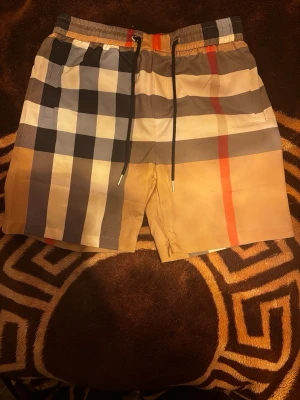 Rutig shorts med snörning Burberry - Säljer ett par klassiska rutiga shorts från Burberry med beige, svart, vit och röd färg. Shortsen har elastisk midja med svart snörning och är tillverkade i ett lätt och bekvämt material. Perfekta för sommaren och ger en stilren look.