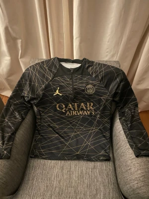 Svart PSG träningsjacka Jordan - Säljer en svart långärmad tränings tröja från PSG och Jordan med guldiga detaljer och loggor. Tröjan har ett coolt geometriskt mönster i guld, halv dragkedja framtill och Qatar Airways tryck på bröstet. Perfekt för fotbollsträning eller chill. I strl xs-s 