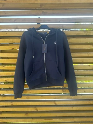 Marinblå zip hoodie från Polo Ralph Lauren - Snygg marinblå hoodie från Polo Ralph Lauren med dragkedja framtill, två stora fickor och klassisk huva med dragsko. Ikoniska vita loggan broderad på bröstet. Tillverkad i mjukt bomullsmaterial som känns skönt mot huden. Perfekt för en avslappnad och stilren look.