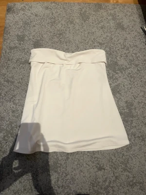 Vit axelbandslös tubtopp - Säljer en stilren vit tubtopp med bred kant upptill. Toppen är axelbandslös och har en enkel, clean look som passar till många olika outfits. Perfekt för varma dagar eller som layering-plagg.