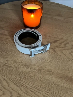 Vit skinnbälte med H-spänne - Säljer ett stilrent vitt bälte i skinn med ett coolt H-format spänne i silver. Perfekt för att lyfta din outfit med en clean och modern touch. Bältet är slätt och har en klassisk design som passar till jeans eller kostymbyxor.