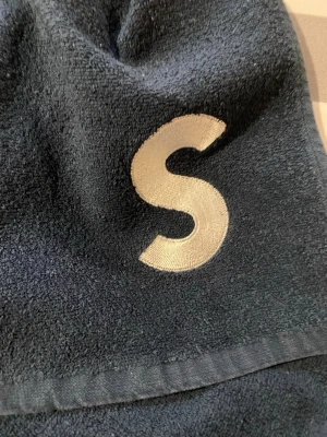 Stilrent SATS blått frotté badlakan med S-brodyr - Mysig blå handduk i frotté med vit broderad bokstav S på ena hörnet. Praktiska hällor på sidorna för enkel användning. Perfekt för att slappa hemma eller efter duschen. Materialet är mjukt och absorberande bomullsfrotté. Marinblått. C:a 60x120 cm. Upptvättad och struken. 