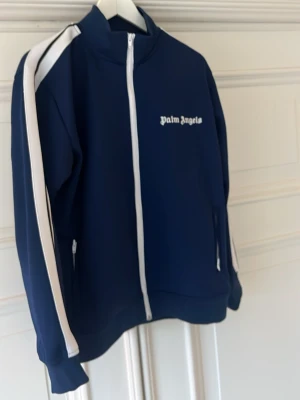 Palm angels tröja  - Snygg marinblå track jacket från Palm Angels med vita ränder längs ärmarna och vit dragkedja. Jackan har hög krage och logga på bröstet. Perfekt för en sportig och trendig look.