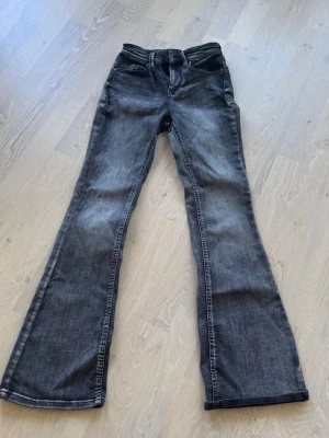 Grå bootcut jeans med hög midja - Säljer ett par grå bootcut jeans med hög midja och klassiska fem fickor. Jeansen har en snygg tvättad look och vida ben som ger en retrovibe. Perfekta för dig som vill ha en avslappnad men trendig stil. Materialet är denim.