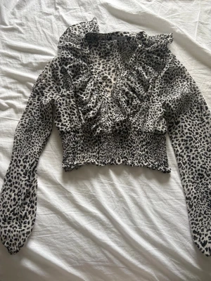 Leopardmönstrad blus med volanger - Snygg blus från Zara med svartvitt leopardmönster, djup V-ringning och volangdetaljer framtill. Blusen har långa ärmar i tunn, luftig kvalitet och smockad midja för en figurnära passform. Perfekt för dig som gillar trendiga statement-plagg.