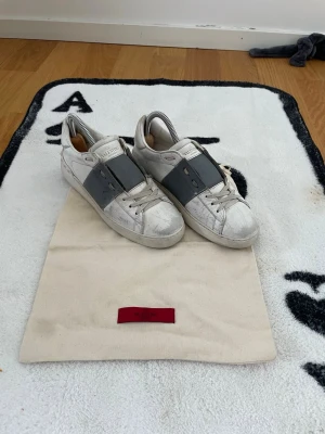 Valentino vita sneakers med grå detalj - Snygga vita sneakers från Valentino. De är väldigt slitna men funkar. Säljer dem för de blivit försmå. Dust bag följer med, ingen box. 24cm mått