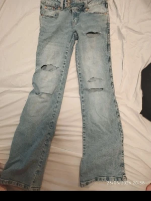 Ljusblå slitna bootcut jeans - Säljer ett par ljusblå bootcut jeans med flera slitningar och hål på benen. Jeansen har klassisk femficksdesign, knappgylf och är tillverkade i denim. Perfekta för en avslappnad och trendig look.