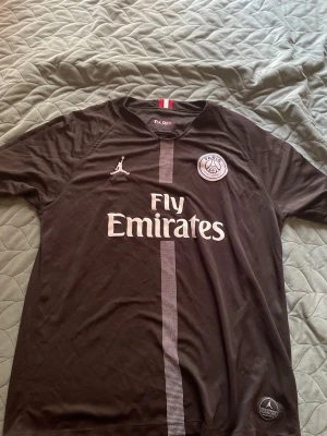 Svart PSG matchtröja Jordan x Fly Emirates - Snygg svart matchtröja från PSG i samarbete med Jordan och Fly Emirates. Tröjan har korta ärmar, rund hals och en vertikal rand i mitten. PSG-logga och Jordan-logga på bröstet, samt Fly Emirates-tryck framtill. Perfekt för fotboll eller streetwear.Storlek Xl
