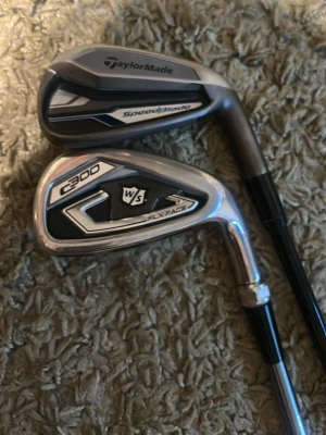Två golfklubbor TaylorMade & Wilson järn 7 or - Säljer två stycken järnsjua golfklubbor, en TaylorMade SpeedBlade och en Wilson Staff C300 FLX Face. Båda klubborna har svart och silverfärgad design med detaljer i metall. Perfekt för dig som vill testa olika märken på banan.