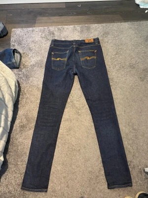Mörkblå skinny jeans från Nudie - Snygga mörkblå skinny jeans från Nudie med klassiska orange sömmar och broderade bakfickor. Jeansen har fem fickor och är tillverkade i stretchig denim för en tight passform. Perfekta för en stilren och modern look.