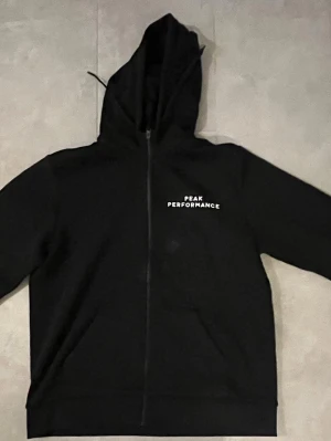 Svart hoodie Peak Performance - Svart hoodie med dragkedja från Peak Performance. Klassisk design med huva och justerbara snören, samt vit logga på bröste och på armen..  passar en större L
