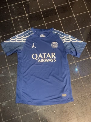 Blå PSG matchtröja Jordan x Qatar - Säljer en blå Paris Saint-Germain matchtröja från Jordan med Qatar Airways-logga på bröstet. Tröjan har vita detaljer och mönster som liknar vingar på axlarna. Materialet är lätt och andas, perfekt för sport och träning.