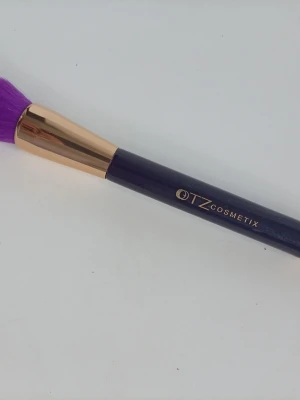Lila sminkborste från TZ Cosmetix - Snygg sminkborste från TZ Cosmetix med lila, täta syntetborst och svart handtag. Metallhylsan är i guldton och ger ett lyxigt intryck. Perfekt för applicering av puder, rouge eller bronzer. Borsten har en rundad form för enkel och jämn applicering.