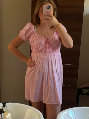 Rosa kort puffärmad klänning - Söt rosa kortklänning med puffärmar och smock i midjan. Klänningen har en rundad urringning med knyt framtill och är tillverkad i ett mjukt bomullsliknande material. Perfekt för en somrig look med voluminösa ärmar och avslappnad passform.