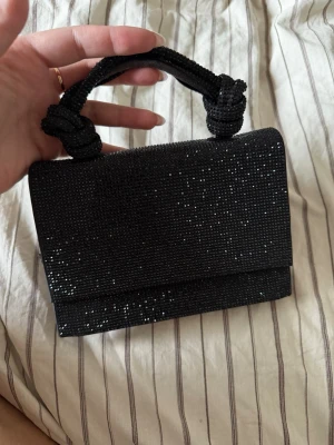 Svart glittrig kuvertväska med handtag - Snygg svart kuvertväska med glittrande pärlor och ett flätat handtag med knutdetaljer. Väskan har magnetlås och ett stilrent innerfack. Perfekt för att lyfta din outfit med lite extra bling. En skada på handtaget som syns på bild, från Zara.