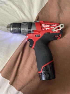 Milwaukee M12 CDD borrmaskin - Kraftfull och kompakt borrmaskin från Milwaukee, modell M12 CDD. Maskinen är röd och svart med ergonomiskt handtag i gummi och metallchuck. Utrustad med LED-lampa och metallclips för bälte. Perfekt för både hemmafix och mer avancerade projekt.