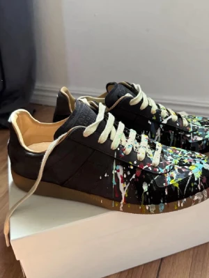 Svarta sneakers med färgstänk - Unika svarta sneakers med färgstänk i vitt, gult, blått och rött. Skorna har beige snörning och innersula samt brun gummisula. Tillverkade i läder med en klassisk låg siluett och cool konstnärlig vibe. Perfekta för dig som vill sticka ut med din stil. Kvitto finns