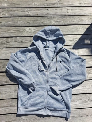 Grå zip hoodie från Polo Ralph Lauren - Säljer en klassisk grå zip hoodie från Polo Ralph Lauren med huva och dragsko. Hoodien har fickor framtill och den ikoniska lilla broderade loggan på bröstet. Tillverkad i mjukt sweatshirtmaterial, perfekt för chill dagar.