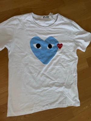 CDG Comme des Garçons Play T-Shirt - Vit CDG T shirt med stort ljusblått hjärta med ett litet rött brevid. Perfekt till sommaren för en riktigt stilig look, XS/S den är i strl S men är något mindre så typ XS