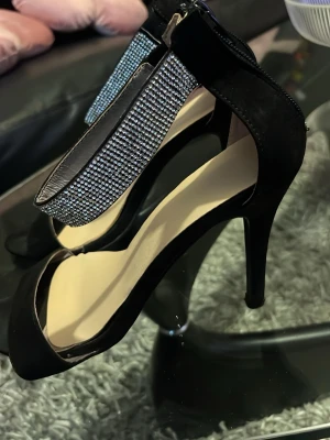 Svarta pumps med strassdetaljer - Snygga svarta pumps med öppen tå och höga smala klackar. Skorna har en bred rem över tårna och en T-rem dekorerad med glittrande strass som ger extra bling. Dragkedja baktill för enkel påtagning. Perfekta för att lyfta din outfit.
