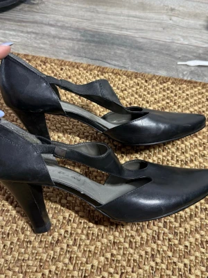Svarta pumps med rem från Tamaris (37) - Säljer ett par svarta pumps från Tamaris i skinn med spetsig tå och bred klack. Skorna har en snygg remdetalj över foten och öppen design vid sidorna. Perfekta för dig som gillar klassisk men trendig stil.