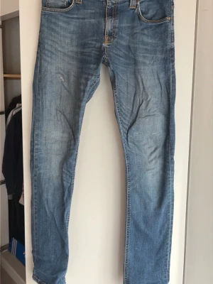 Blå vintige jeans från Nudie Jeans - Säljer ett par klassiska blå vintige jeans från Nudie Jeans med fem fickor och snygga slitningar. Jeansen har en rak passform och är tillverkade i mjuk denim. Bakfickorna har Nudie Jeans karaktäristiska broderi i orange. Perfekta för en avslappnad stil.