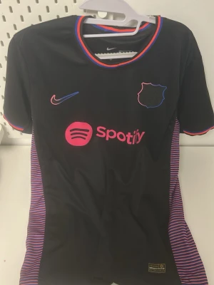 Svart Nike FC Barcelona tröja - Snygg svart FC Barcelona matchtröja från Nike med Spotify-logga i rosa på bröstet. Tröjan har färgglada detaljer i blått, rött och rosa runt kragen och ärmsluten samt randiga sidopaneler. Tillverkad i lätt och ventilerande material, perfekt för träning eller match.