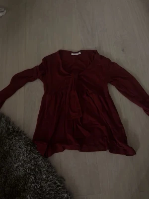 Vinröd blus med knyt framtill - Snygg vinröd blus med långa ärmar och knytning framtill. Blusen har en lös passform och ett mjukt, följsamt tyg som ger en avslappnad men trendig look. Perfekt att styla med jeans eller kjol för en cool vardagsstil.
