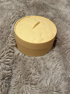 Gul rund förvaringsburk i plast äkta till Dumplinh - Säljer en rund förvaringsburk i gul plast med lock. Locket har ett flätat mönster och ett litet handtag på toppen. Perfekt för småprylar, smycken eller andra småsaker. Enkel och stilren design som passar i de flesta rum.