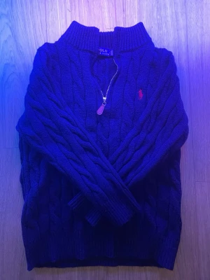 Ralph Lauren Tröja - Mörkblå kabelstickad tröja från Polo Ralph Lauren med halv dragkedja och röd broderad logga på bröstet. Tröjan har hög krage och ribbade muddar. Klassisk och tidlös design i mjukt material. Storlek S-M. Har med tagen, användes 1-2 gånger.