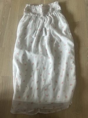 Vit långkjol med små rosa blommor - Supersöt vit långkjol med små rosa blommor och smock i midjan. Kjolen har knytband och är gjord i ett lätt, luftigt tyg som passar perfekt till varmare dagar. Den har en romantisk och feminin känsla med volangliknande fall.